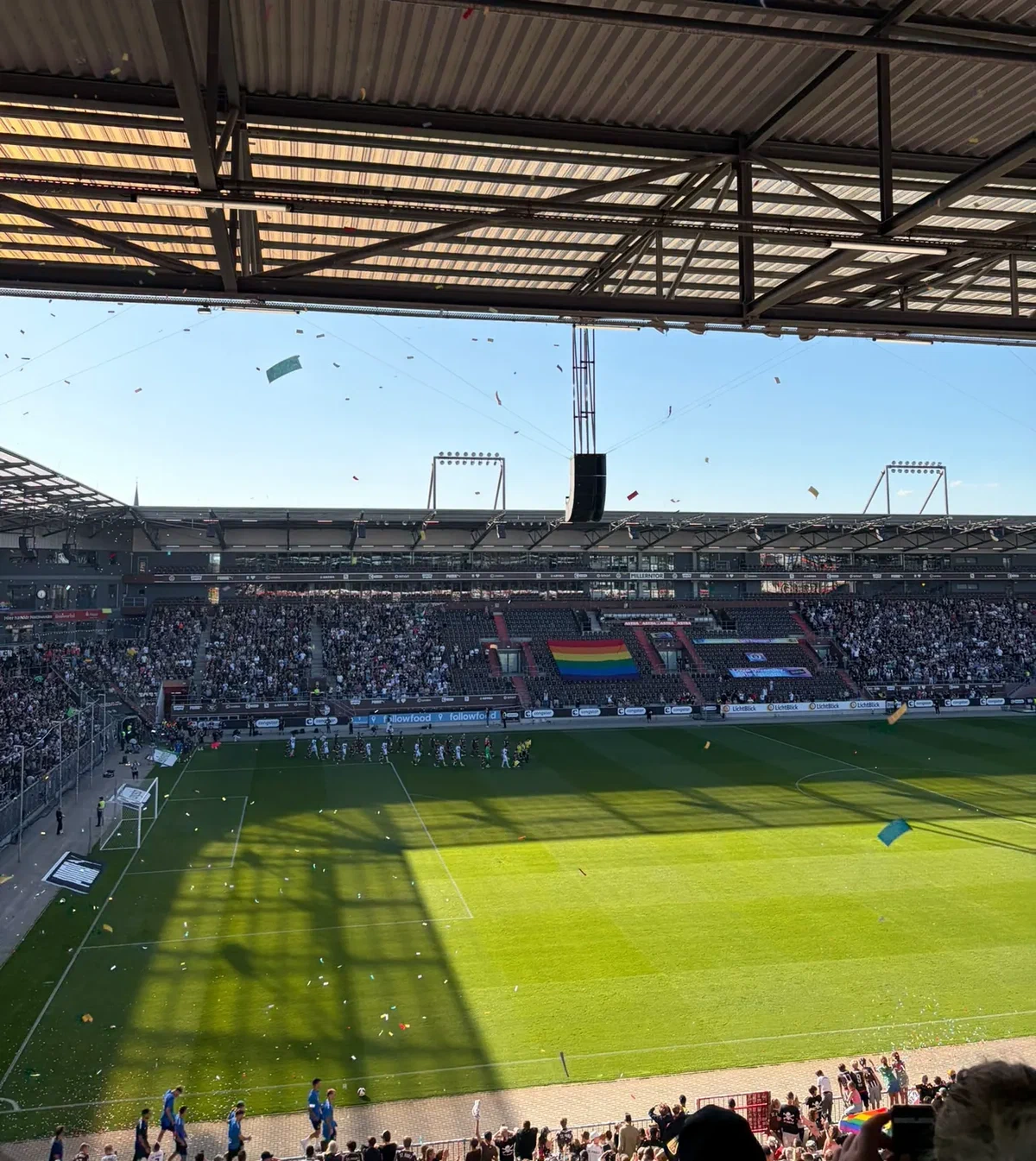 Blick von der Gegengerade auf den Rasen des Millerntor-Stadions, Konfetti in der Luft und eine Regenbogenfahne auf der Gegentribüne